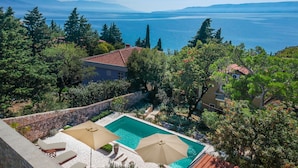 Un espace piscine agrémenté de deux parasols, d’un mur en pierre et offrant une vue sur la mer et les montagnes.