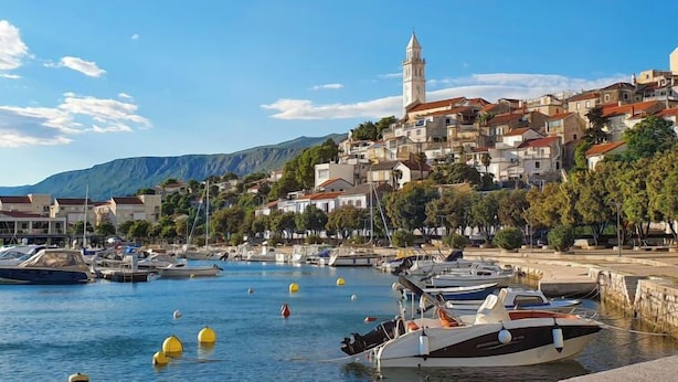 Une ville côtière dotée d’une tour imposante, où des bateaux sont amarrés le long du port, et qui est bordée de collines.