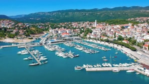 Une ville côtière dotée d’un port de plaisance rempli de bateaux, entourée de bâtiments résidentiels et bordée de montagnes en arrière-plan.