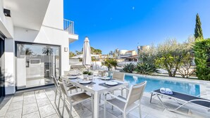 Un área de comedor exterior moderna con piscina, mobiliario blanco y vistas despejadas del vecindario.