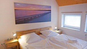 A bedroom with a large bed, wooden headboard, bedside lamps, a painting of a sunset, and a window with curtains. - Vacation home Reuse by Interhome (Norddeich)