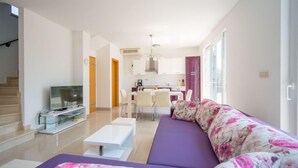 Een moderne woonkamer met een bank, een salontafel en een televisie. Achtergrond is een keuken met een eethoek.