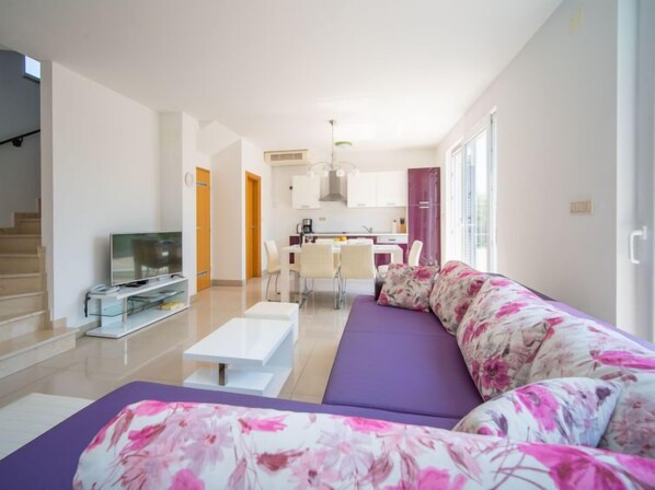 A modern living room with a sofa, a coffee table, and a TV. There is a kitchen in the background with a dining area.