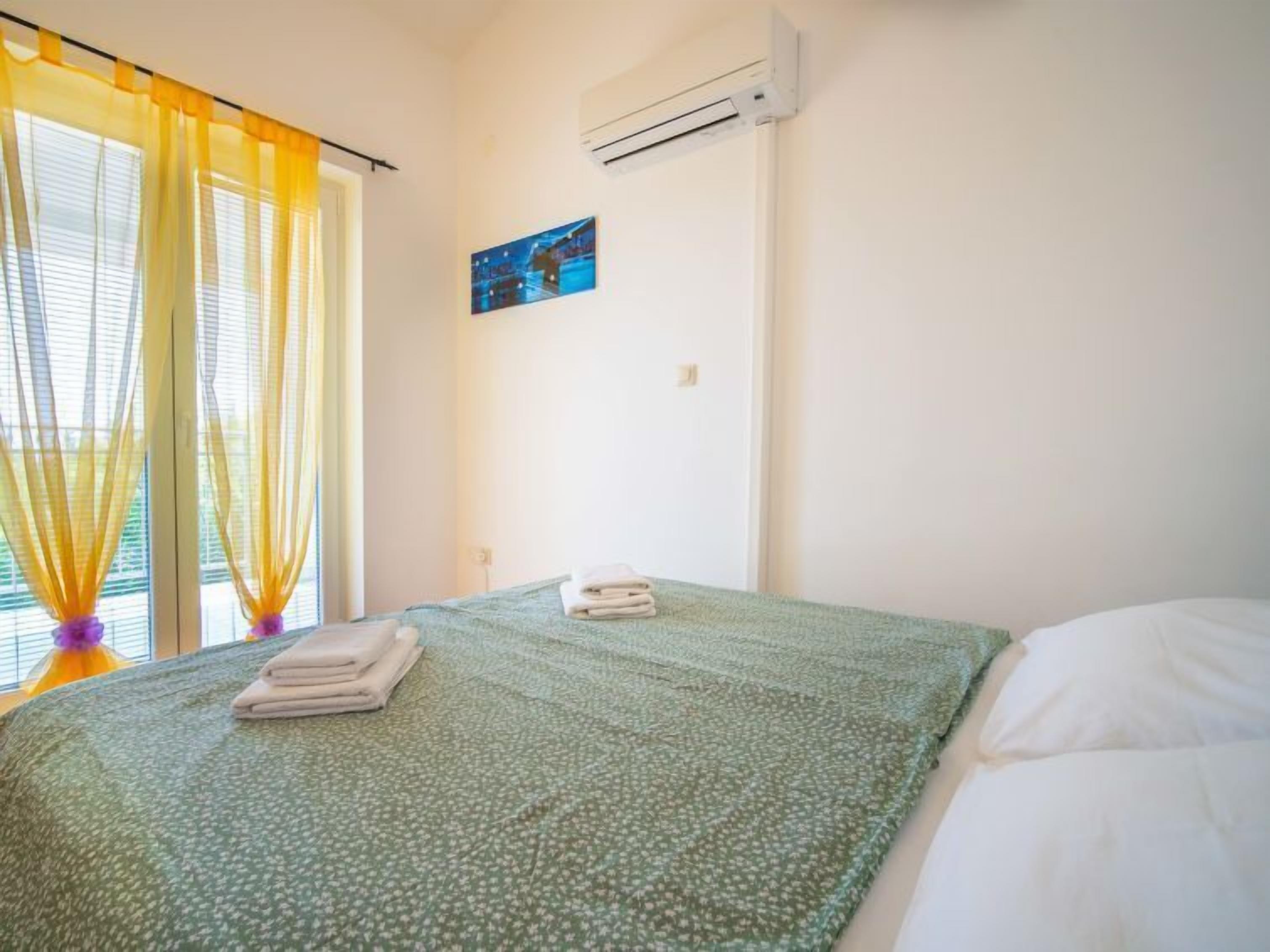 Una habitación de hotel con una cama, una ventana con cortinas amarillas, un equipo de aire acondicionado y un pequeño cuadro en la pared.