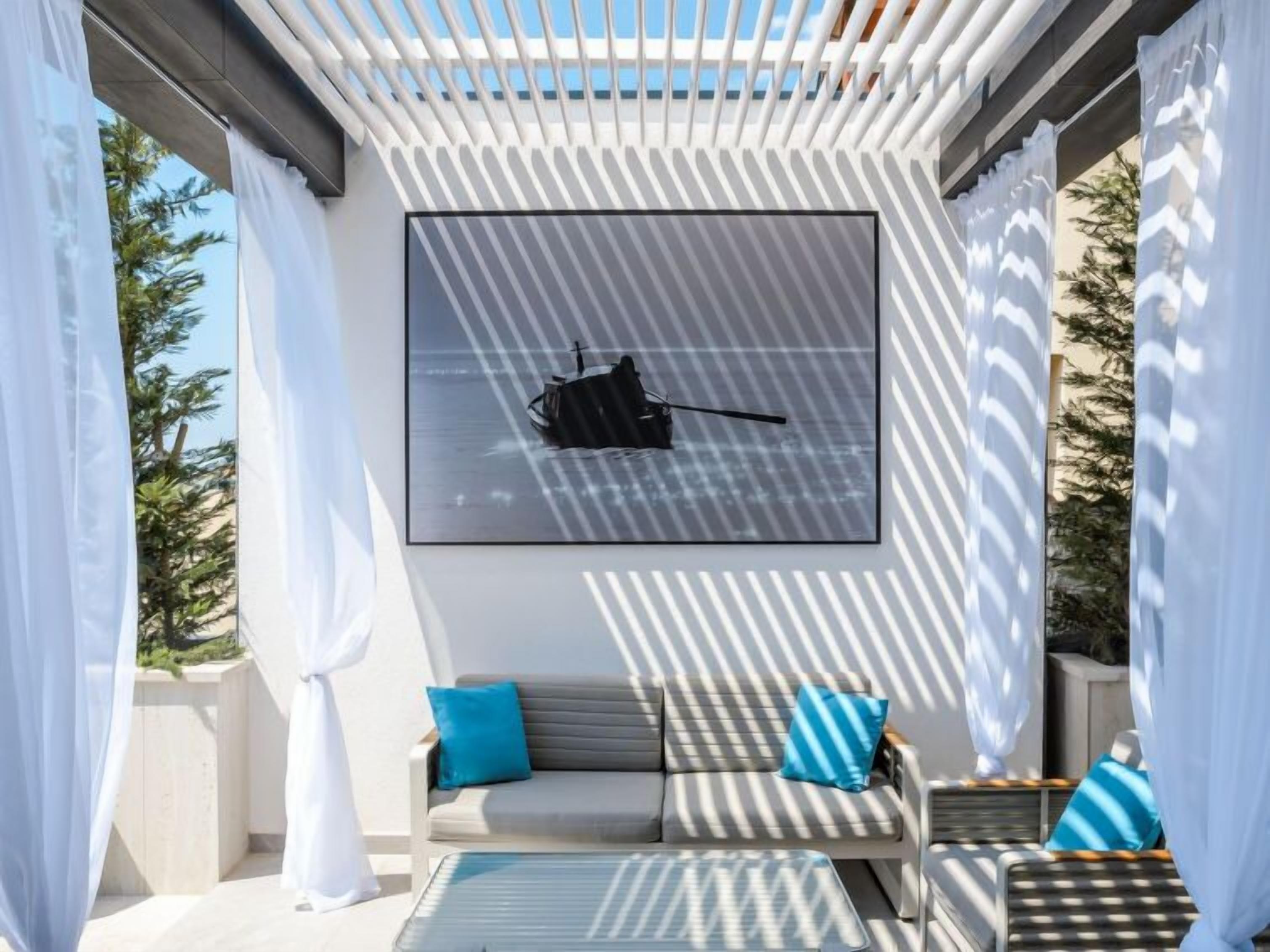 Een zonnig terras met een bank en stoelen, een groot fotokader aan de muur en witte gordijnen die de ruimte omlijsten.