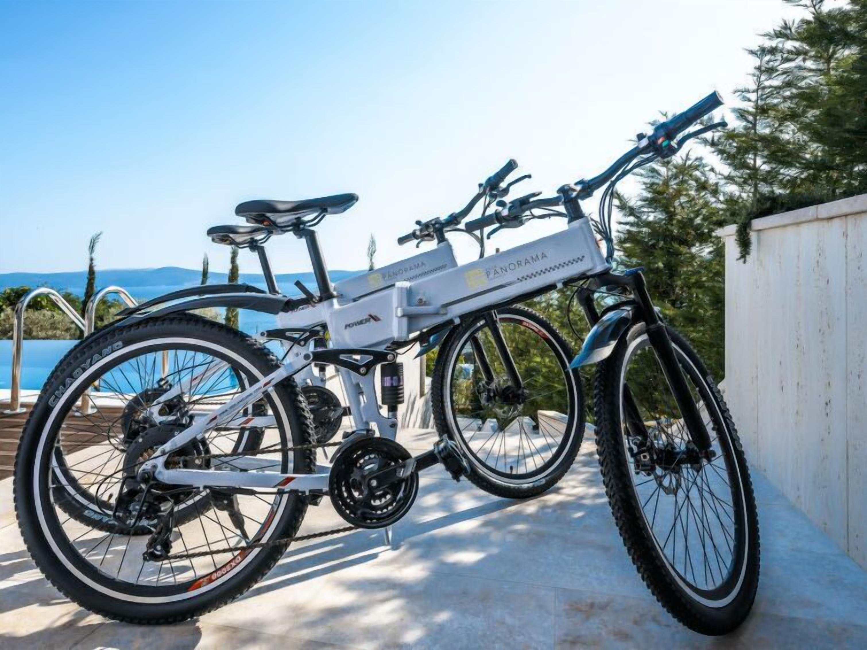 A stationary electric bike with a white frame and black components, parked outdoors with a view of a pool and mountains in the background.