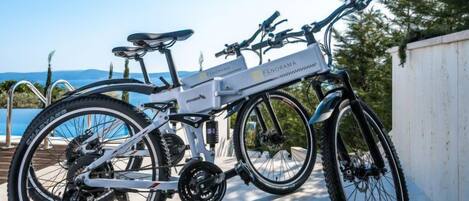A stationary electric bike with a white frame and black components, parked outdoors with a view of a pool and mountains in the background.