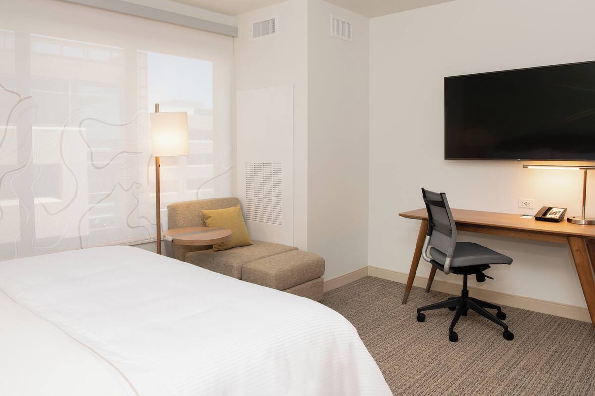 A hotel room with a bed, a desk with a chair, a television, and a lamp.
