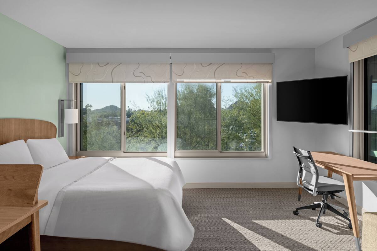 A hotel room with a bed, desk, chair, TV, and large windows offering a view of trees and hills.
