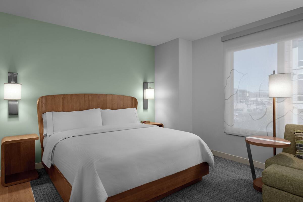 A hotel room with a wooden bed, two bedside tables with lamps, a small round table, and a view of the city through a large window.