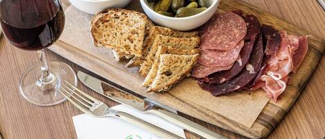 A wooden board with sliced meats, pickles, and bread, accompanied by a glass of red wine and cutlery on a wooden table.