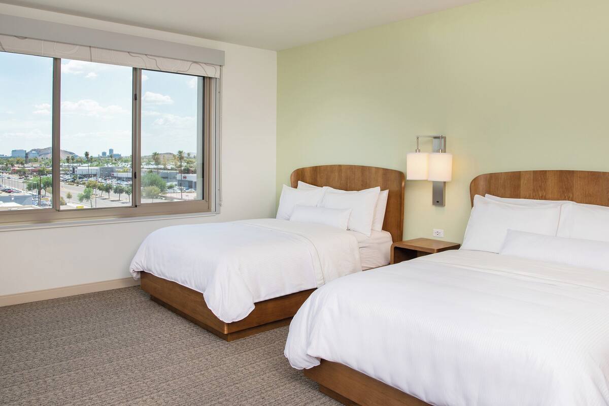A hotel room with two beds, a large window with a city view, and a nightstand with a lamp.
