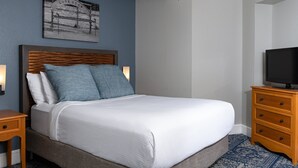 Ein ordentlich bezogenes Bett mit weißen Leinen und blauen Kissen, ein Holz-Kopfteil, ein Nachttisch mit Lampe, eine Holzkommode mit Fernseher und ein Deckenventilator.
