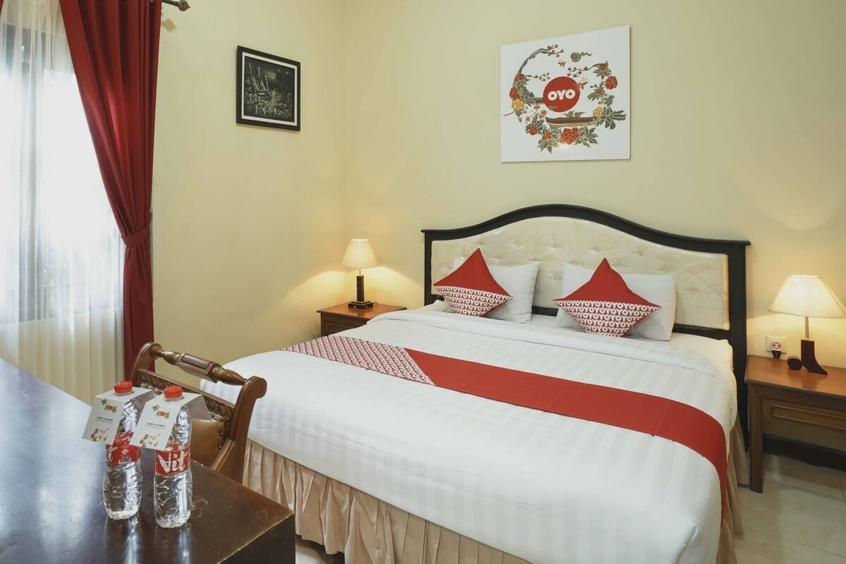Ein Hotelzimmer mit einem ordentlich bezogenen Bett, zwei Nachttischen mit Lampen, einem Esstisch mit Stühlen und einem Fenster mit roten Vorhängen.