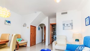 Een woonkamer met een blauwe bank, een eettafel met stoelen, een klein wit tafeltje met een blauwe schaal en een gang die naar een keuken leidt.