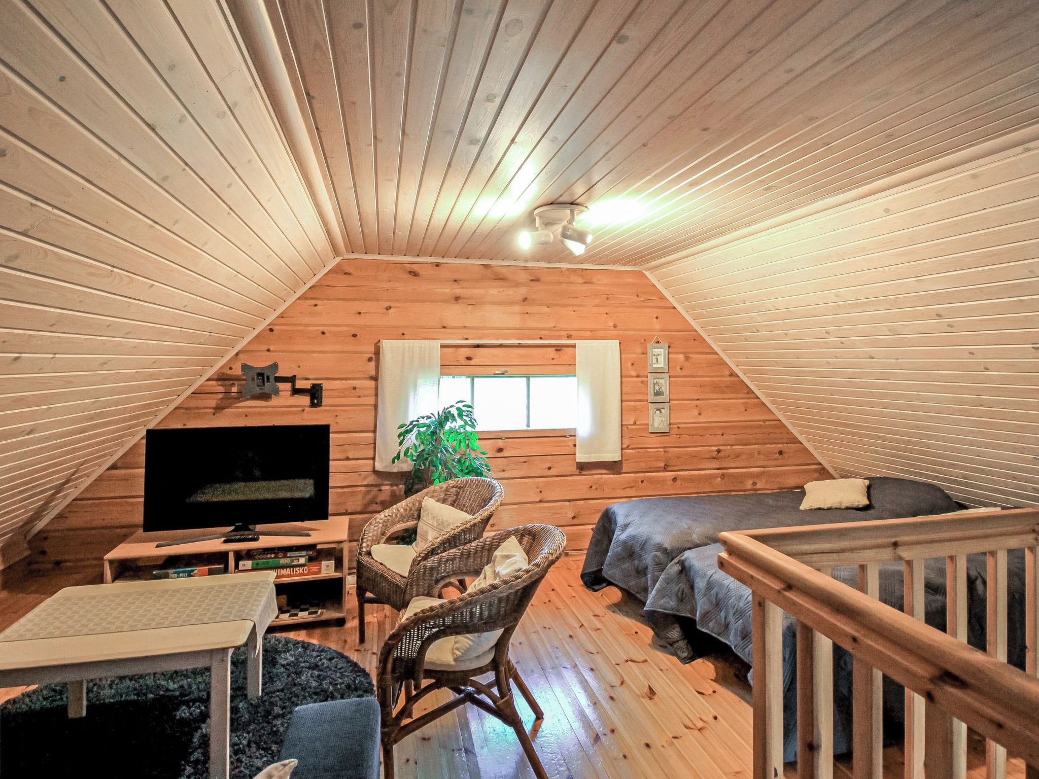 A cozy attic bedroom with wooden walls, a TV, a sofa, a chair, and a window.