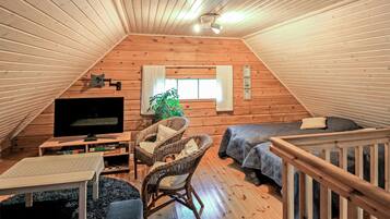 A cozy attic bedroom with wooden walls, a TV, a sofa, a chair, and a window.