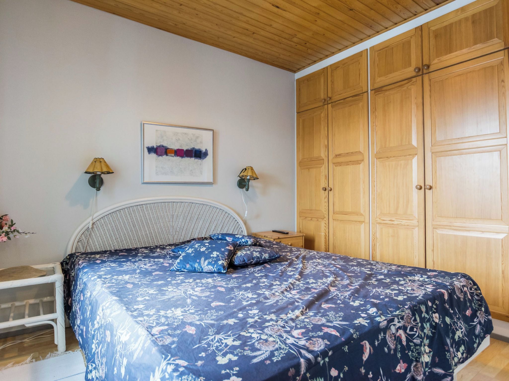 A bedroom with a bed, a nightstand, a wall-mounted lamp, a framed artwork, and wooden wardrobe doors.