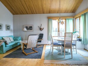 A cozy living room with a wooden ceiling, a teal sofa, a white armchair, a dining table with chairs, and a window with curtains. - Palokärki by Interhome (Kuusamo)