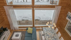 L’intérieur d’un chalet accueillant, doté d’une grande fenêtre donnant sur un paysage enneigé, d’un canapé, d’une table à manger et d’un téléviseur.