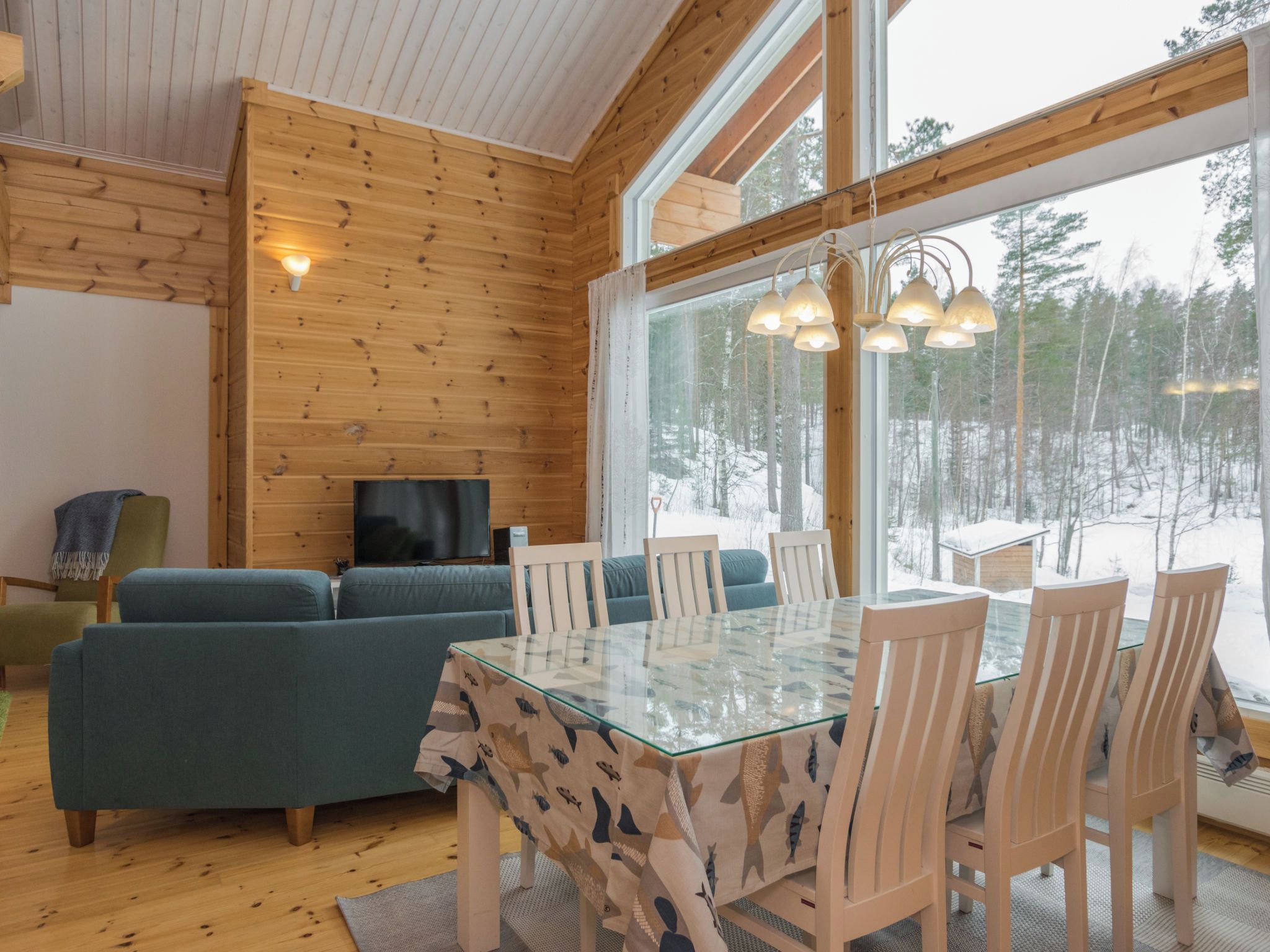 A wooden interior with a glass dining table, chairs, a sofa, and a TV.