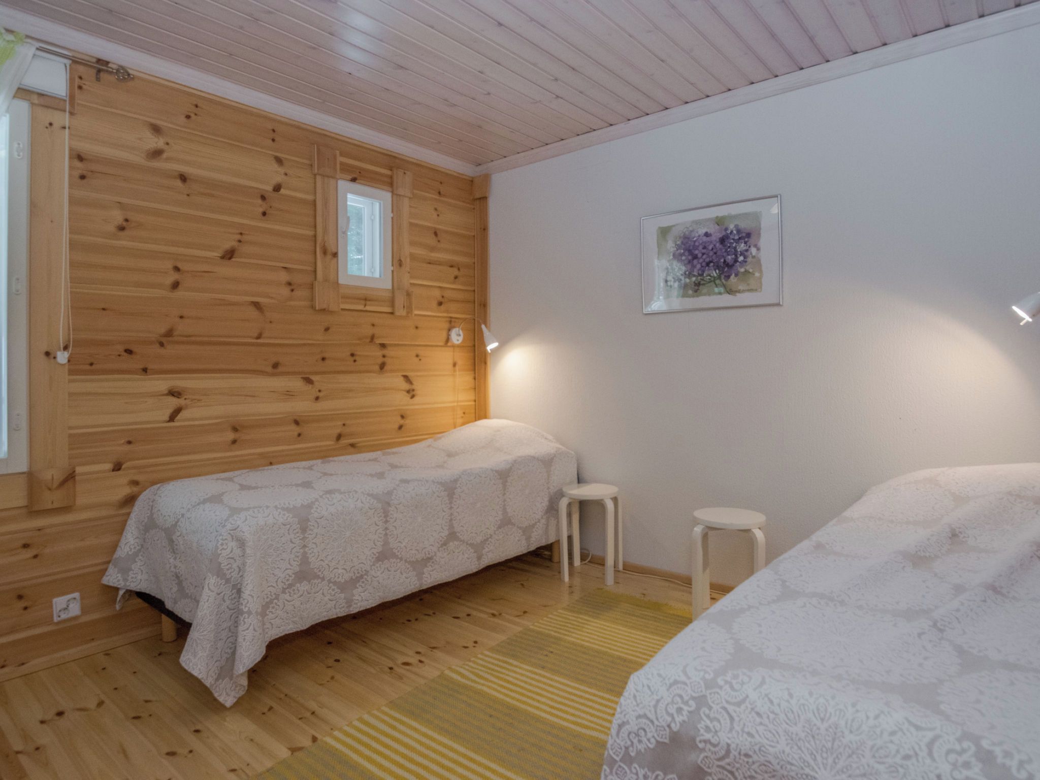 A room with two beds, a small stool, a window, and a framed picture on the wall.