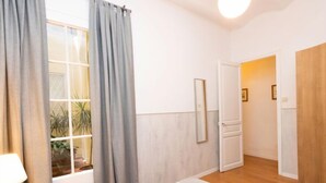 Un dormitorio con una cama, una mesita de noche, una lámpara, una ventana con cortinas, un espejo y una puerta que da a otra habitación.