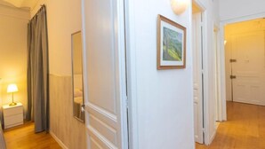 Un pasillo con suelo de madera, paredes blancas, un cuadro enmarcado, un espejo y una puerta que da a otra habitación.