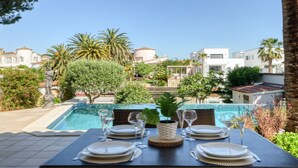 Une table à manger dressée pour quatre personnes, avec vue sur une piscine et des bâtiments en arrière-plan.