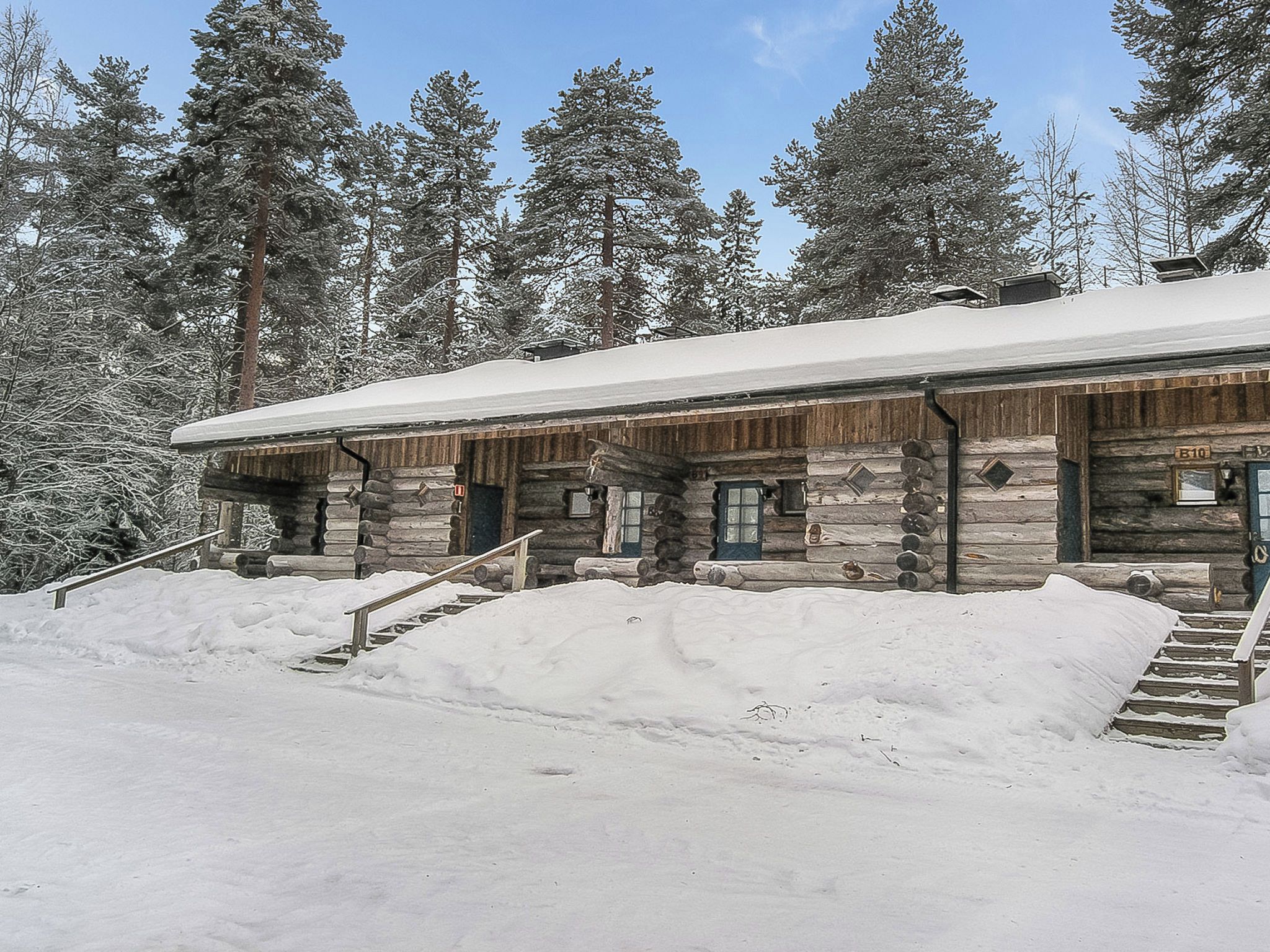 Une cabane en rondins dont le toit est recouvert de neige, avec une façade en bois et un escalier menant à l’entrée.