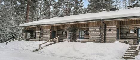 Une cabane en rondins dont le toit est recouvert de neige, avec une façade en bois et un escalier menant à l’entrée.