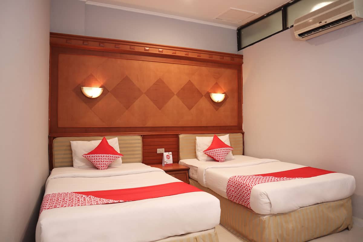 A hotel room with two beds, each with white bedding and red accents, a wooden headboard with wall sconces, and an air conditioning unit on the wall.