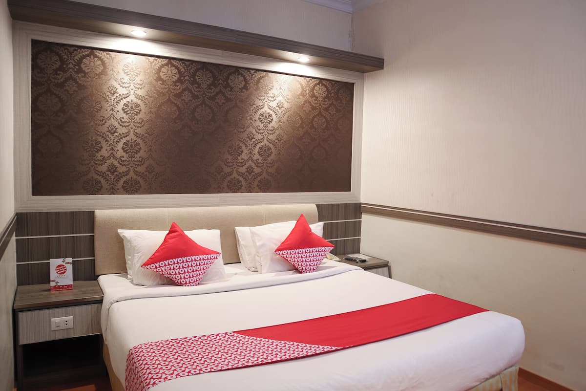 A hotel room with a bed, two pillows, a nightstand, and a red and white patterned blanket.