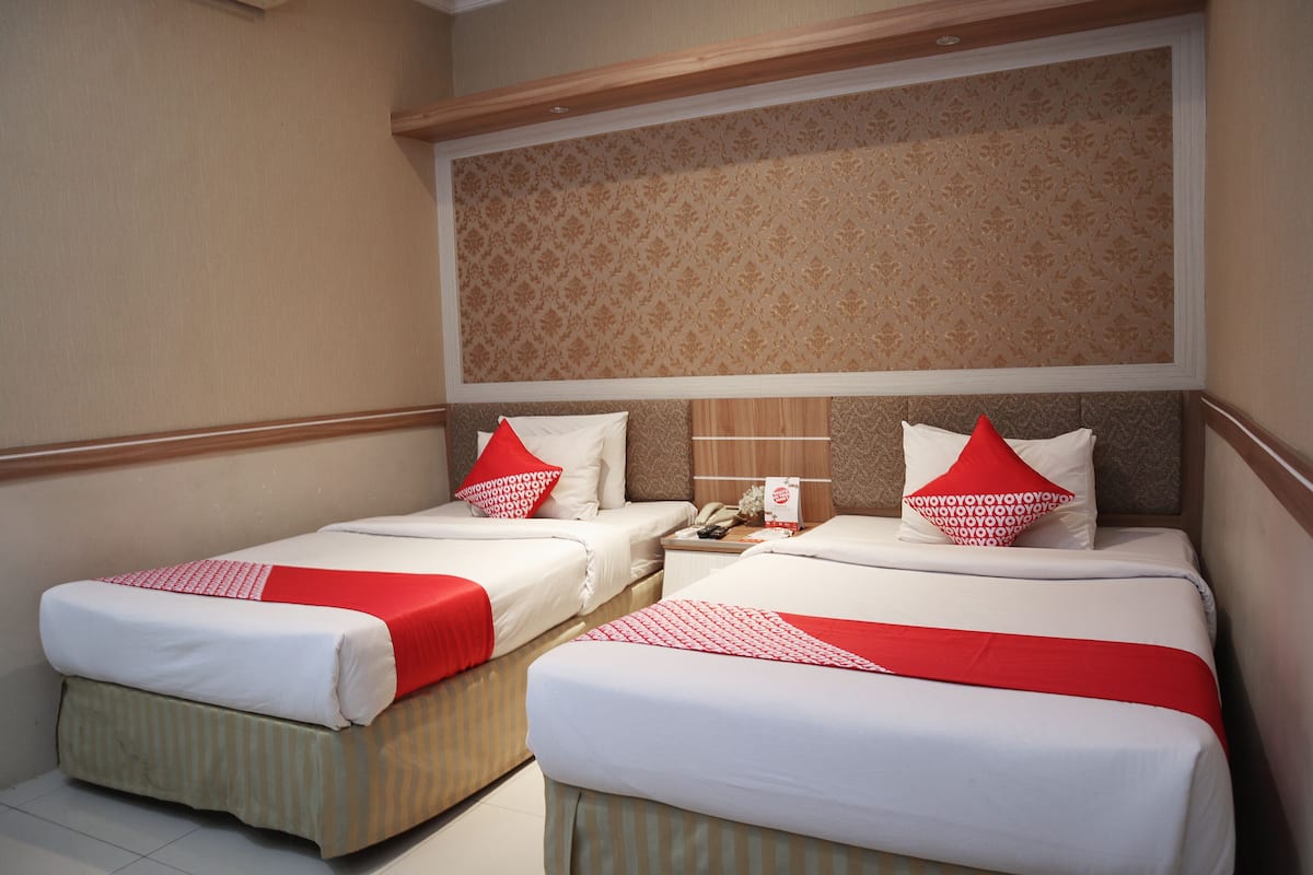 A hotel room with two beds, each with white bedding and red accents, a patterned headboard, and a small bedside table with a lamp.