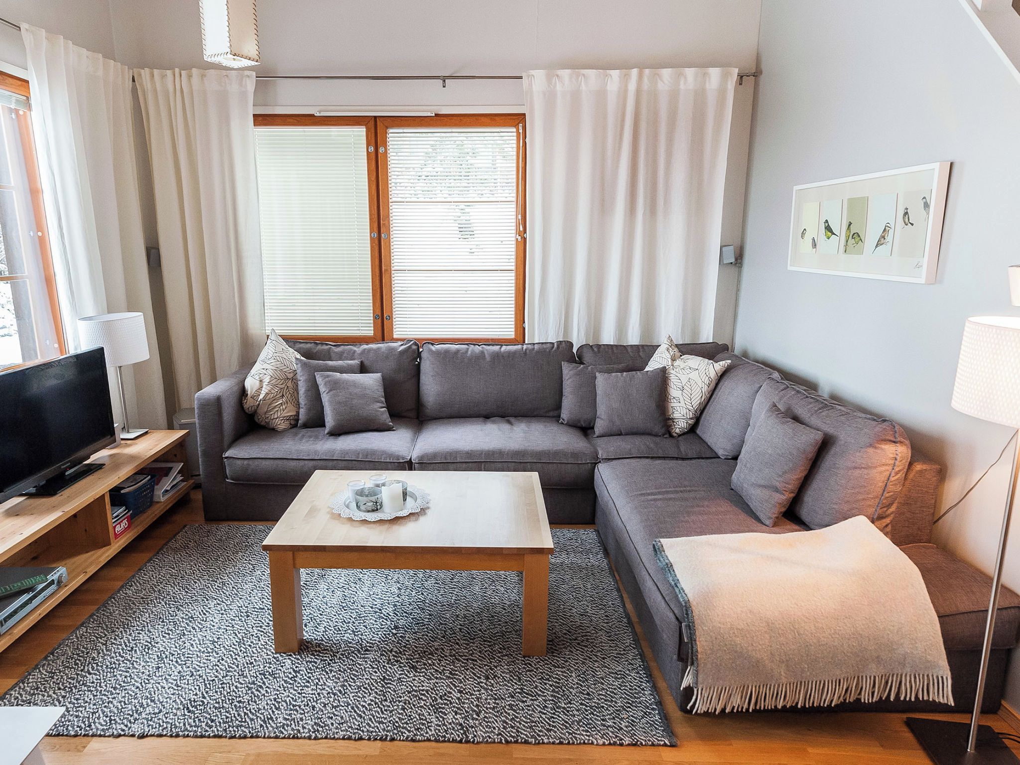 Un salon avec un canapé gris, une table basse, un téléviseur et une fenêtre avec des stores.