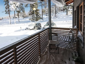 Un balcón cubierto de nieve con baranda de madera, una silla y vista a coches estacionados y árboles.