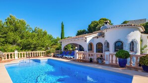 Une piscine avec des chaises longues, une pergola et un bâtiment blanc orné d’arches et surmonté d’un toit en tuiles.