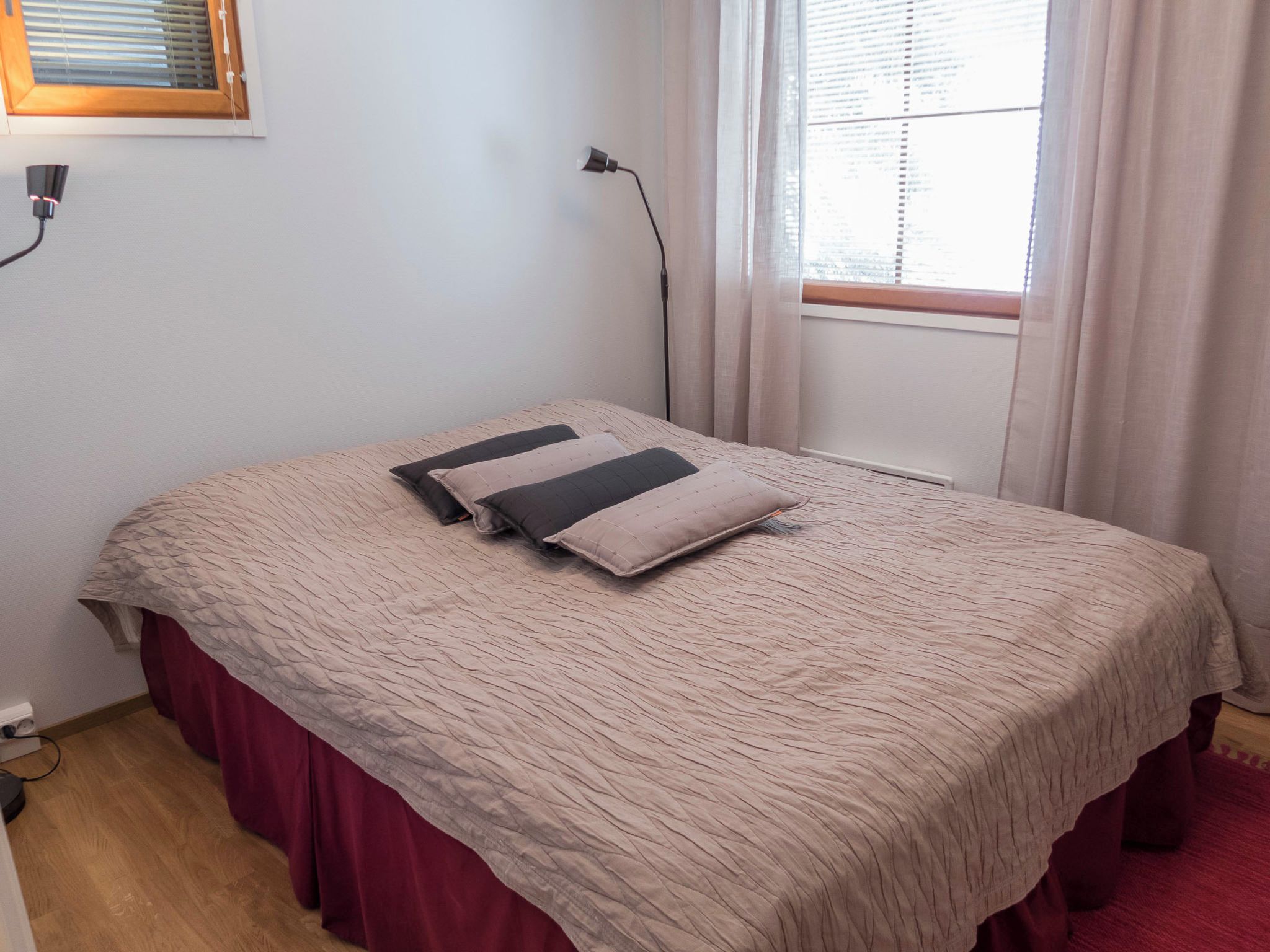 Uma cama de solteiro com colcha bege e duas almofadas, uma janela com cortinas, um abajur e uma tomada na parede.