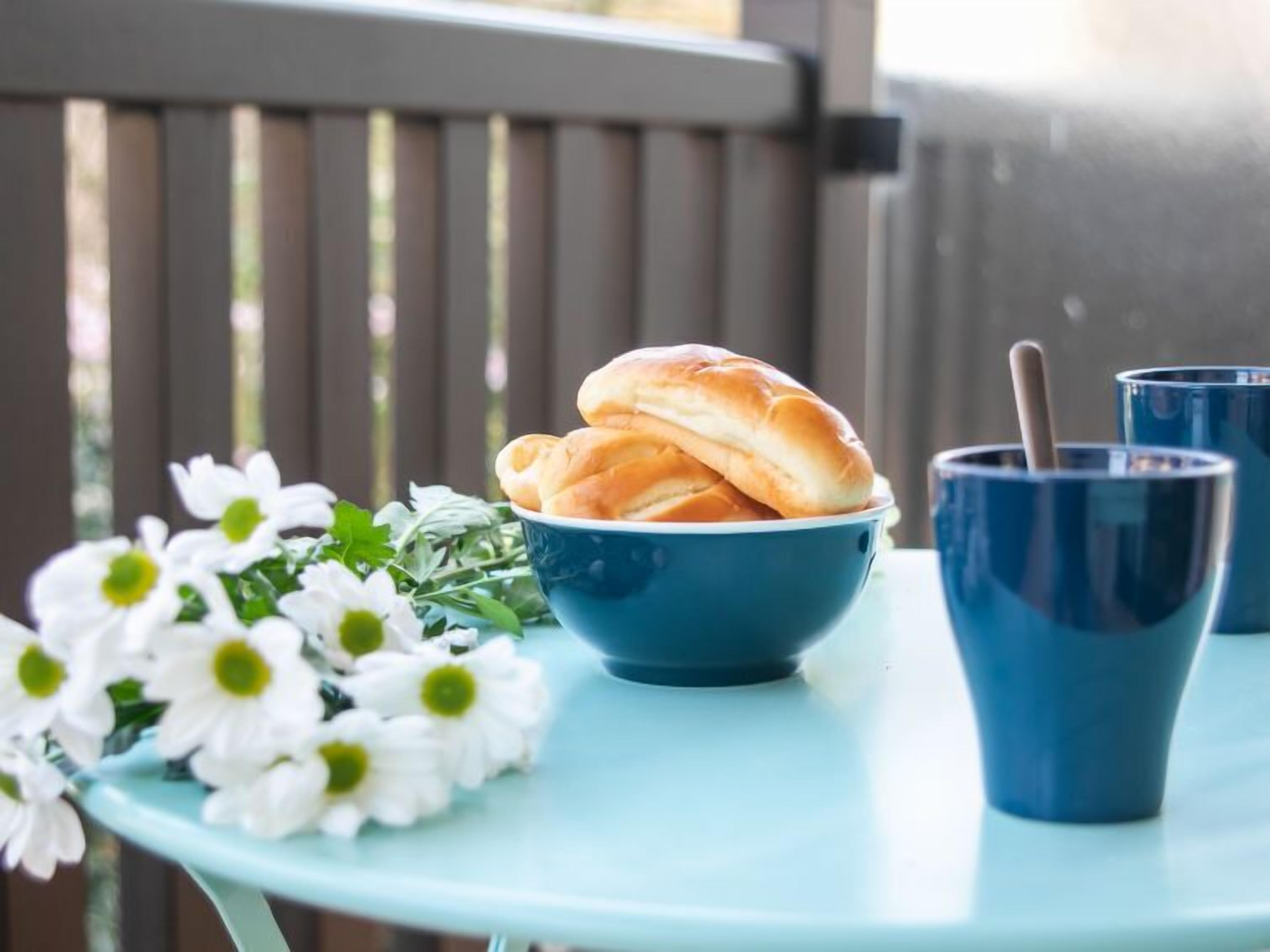 A table with a bowl of bread rolls, a blue mug, and a bouquet of white flowers.