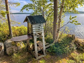 A wooden birdhouse with a sloped roof, situated on a wooden platform with a bench, surrounded by trees and a body of water in the background. - Nanguvilla by Interhome (Inari)