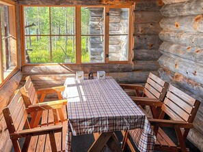 A log cabin interior with a plaid table and benches, large windows with a forest view, and a rustic wooden interior.
