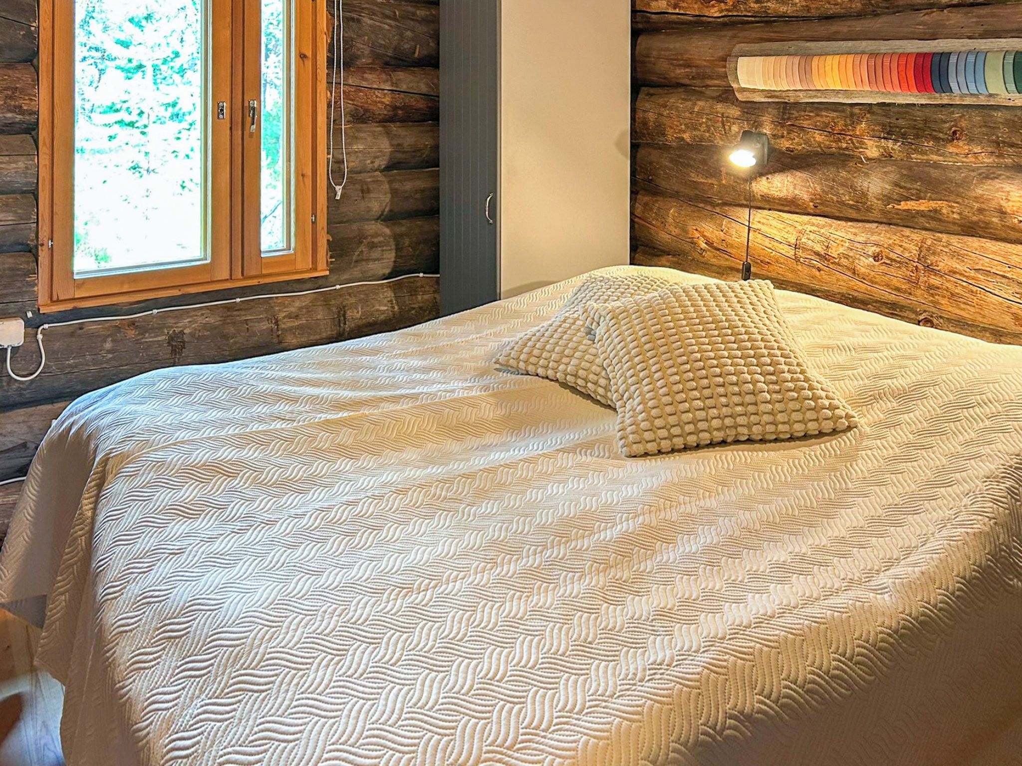 A bedroom with a wooden log wall, a bed with a patterned bedspread, a window with a view, and a wall-mounted light fixture.