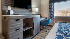 A hotel room with a desk, a flat-screen TV, a blue chair, a bed with a patterned bedspread, and a window with curtains.