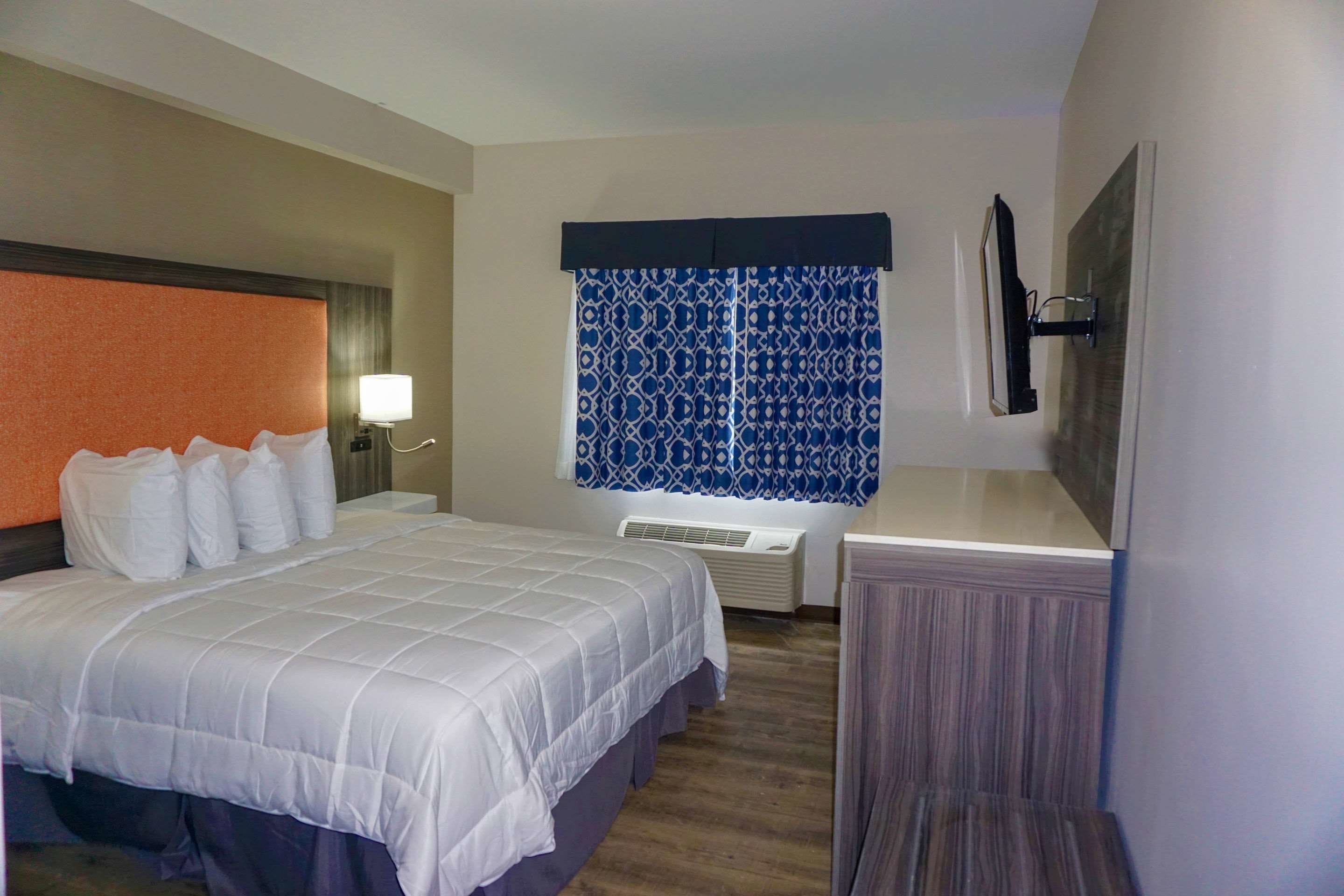 A hotel room with a bed, bedside table, lamp, air conditioning unit, and a window with patterned curtains.