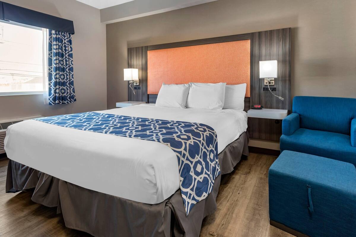 Ein Hotelzimmer mit einem großen Bett, zwei Nachttischen mit Lampen, einem blauen Sessel und Blick ins Freie durch ein Fenster mit gemusterten Vorhängen.