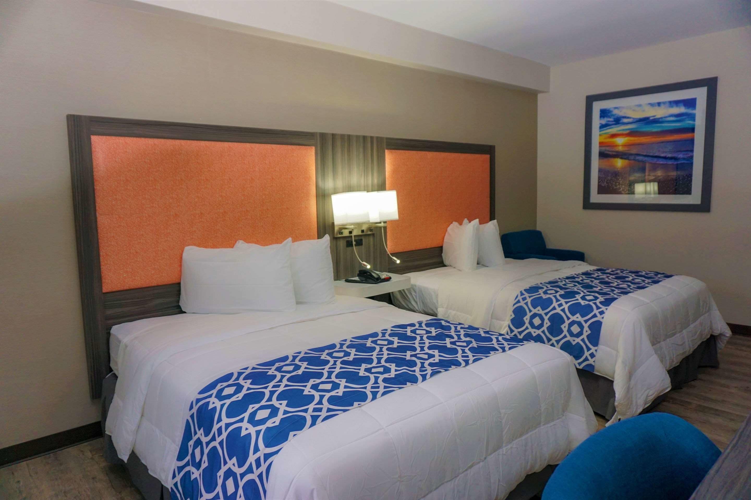 Habitación de hotel con dos camas, una mesita de noche con lámpara, un cuadro en la pared y una silla azul.