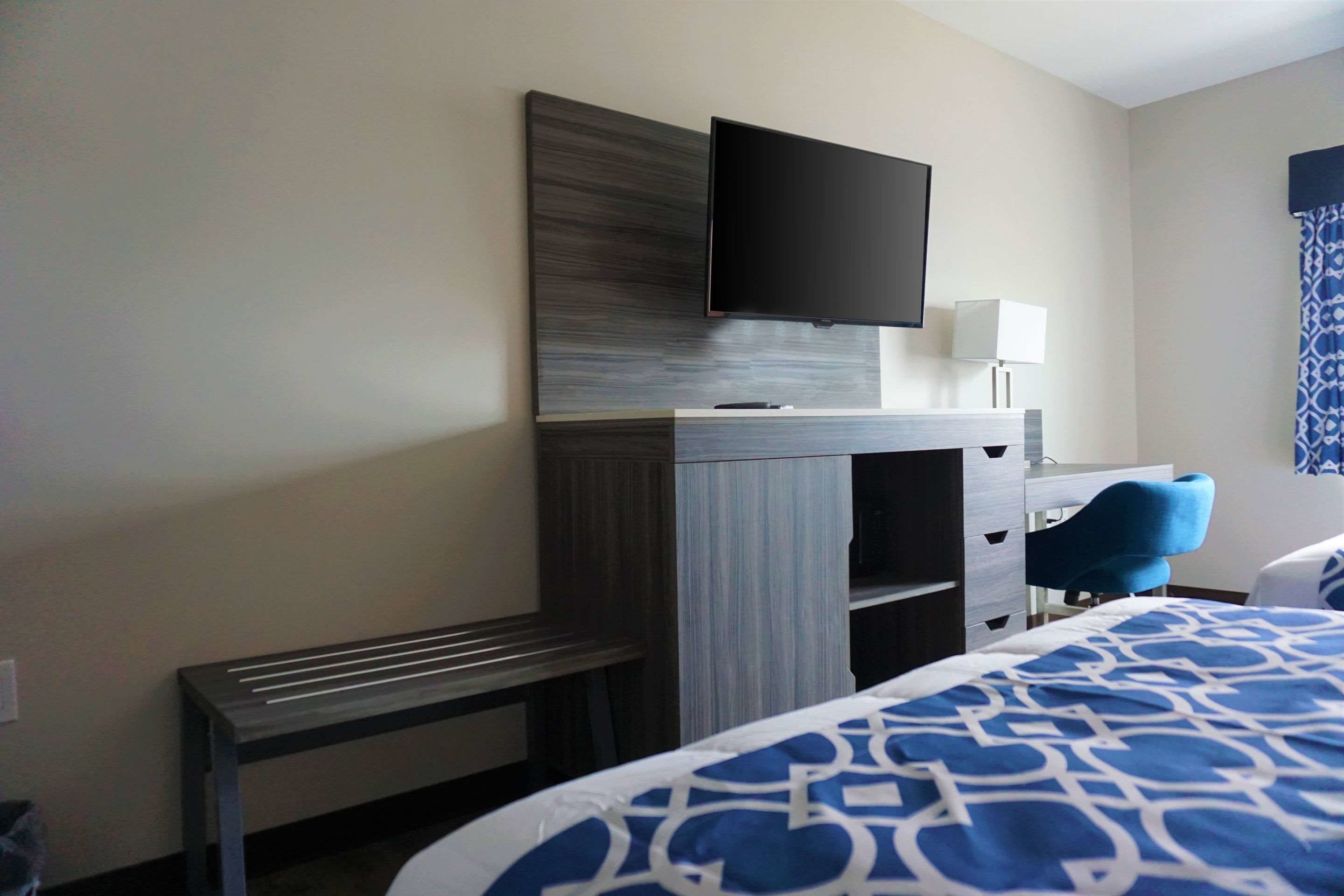 A hotel room with a bed, a desk with a flat-screen TV, a chair, and a lamp.