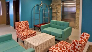 A lobby with teal sofas, patterned armchairs, a woven coffee table, and a metal elevator.