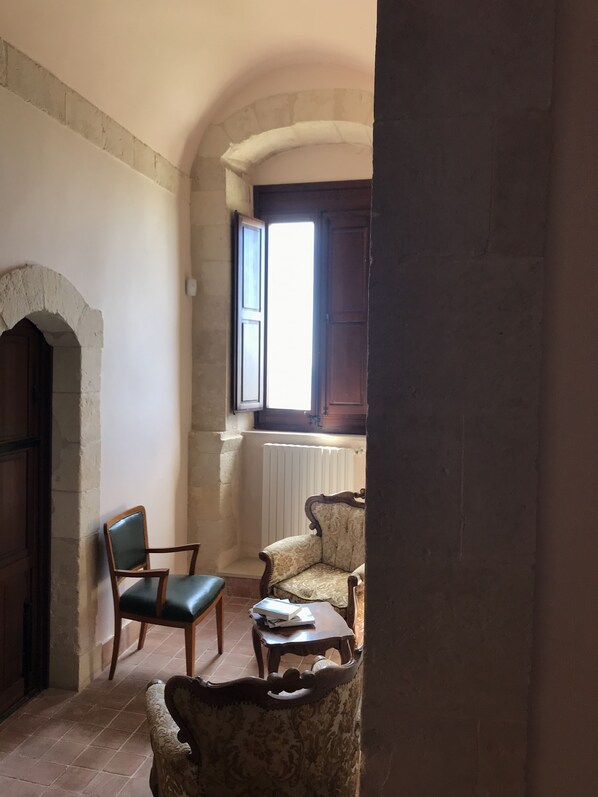 Una habitación con un arco de piedra, una puerta de madera, una ventana con contraventanas de madera y dos sillones tapizados.