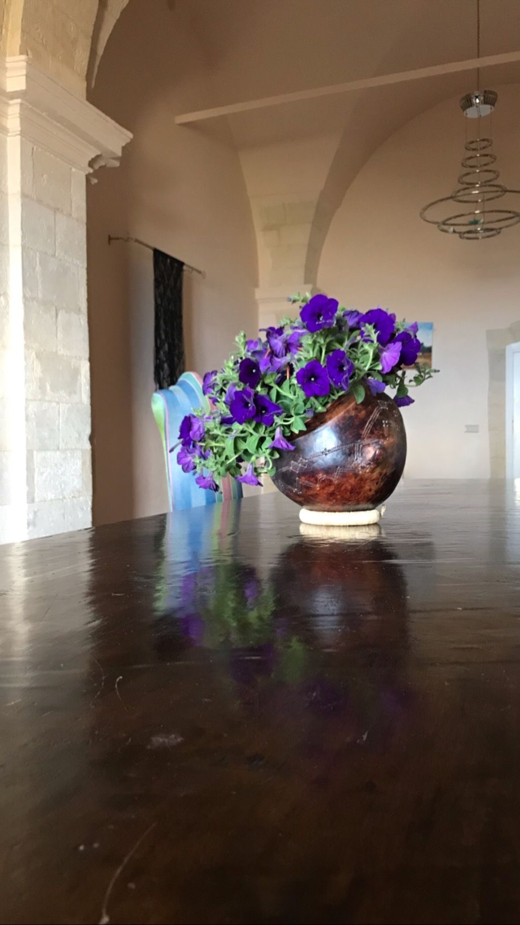 Un jarrón grande y ornamentado con flores moradas sobre un suelo de madera pulida en una habitación amplia con techos altos y una araña.